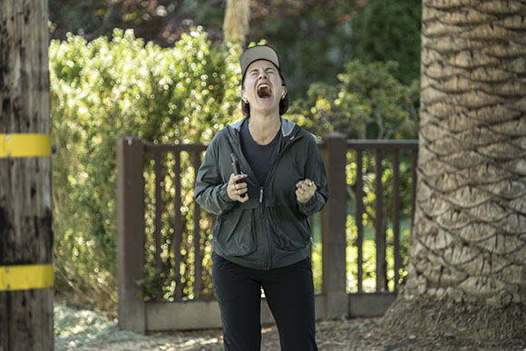 A person in a grey jacket and black cap stands outdoors with their mouth wide open in a shout.