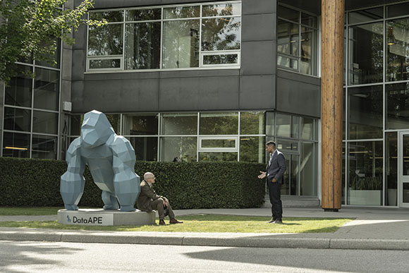 A large blue geometric gorilla sculpture sits on a platform near a man standing and another man sitting on the grass.