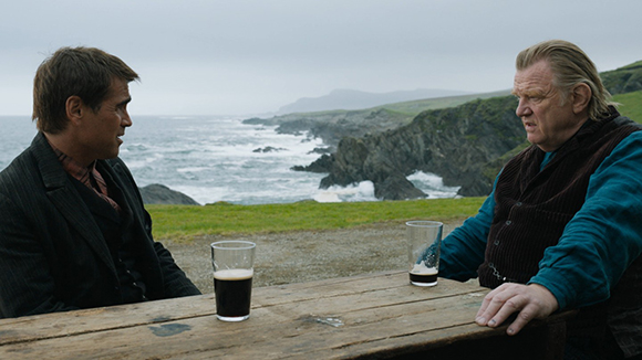 Two men sit at a wooden table with drinks, facing each other against a backdrop of a rocky ocean coast.