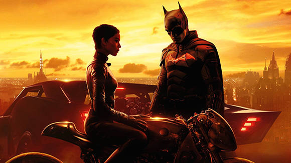 Batman stands beside Catwoman on a motorcycle against a glowing orange city skyline.