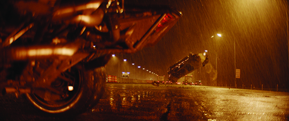 A large vehicle flips over on a rain-slicked road under yellow streetlights at night.
