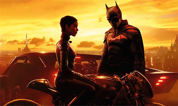Batman stands beside a woman on a motorcycle against a bright orange sunset skyline.