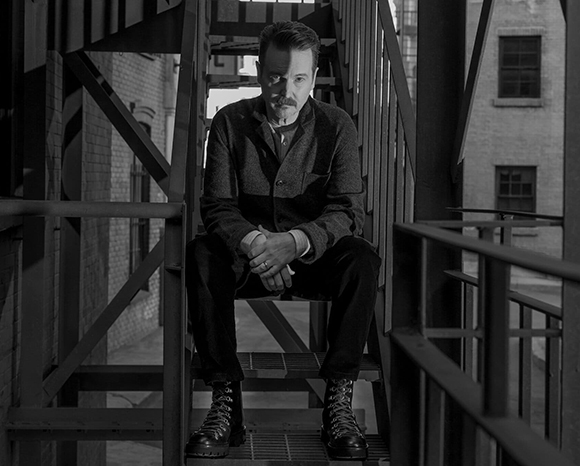 A man sits on metal stairs in a dark, industrial environment with shadows and structural beams.