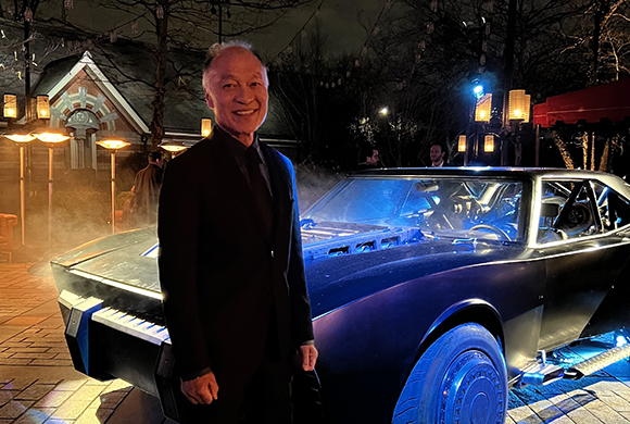 A smiling man in a black suit stands beside a sleek, dark, futuristic car outdoors at night.