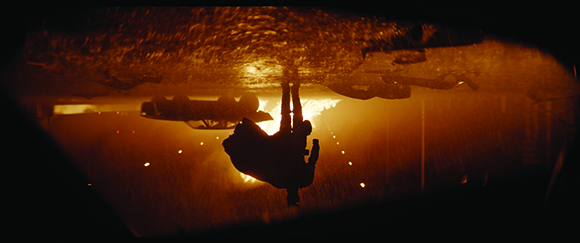 Silhouette of a person hanging upside down in a dark, orange-lit environment with rain or texture.