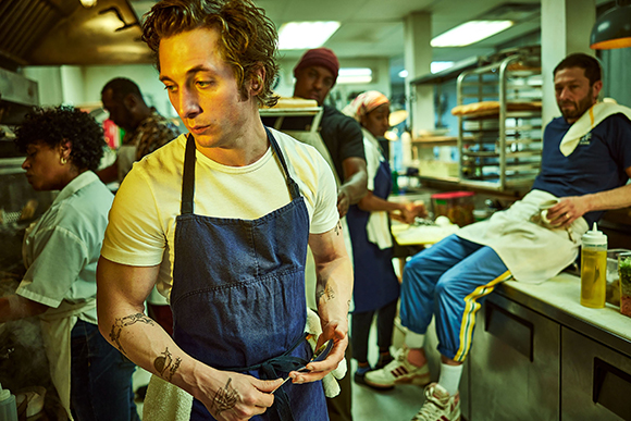 A male chef in a blue apron works in a crowded, brightly lit professional kitchen.