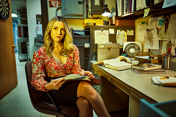 A woman in a floral blouse sits at a desk covered in papers and books in a cramped office.
