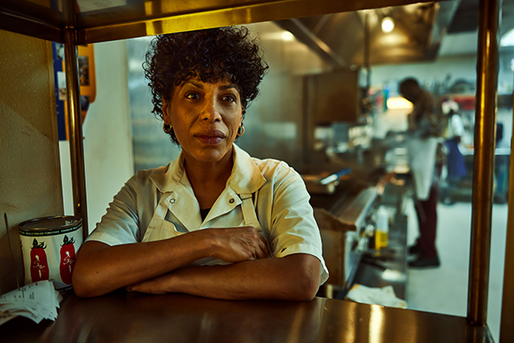 A woman with curly dark hair in a white chef's coat leans on a counter in a professional kitchen.