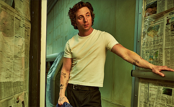 A man with wavy brown hair and tattoos on his arms wears a white t-shirt and dark trousers.