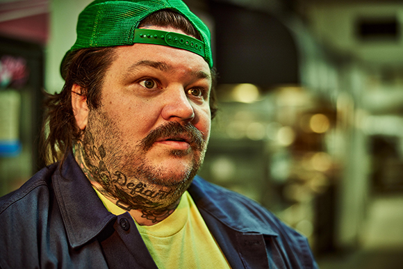 A man with a mustache, tattoos on his neck, a green cap, and a blue jacket looking to the side.