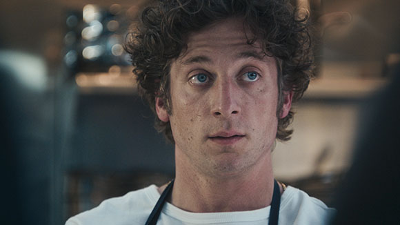 A man with curly brown hair and blue eyes looking forward in a kitchen setting.