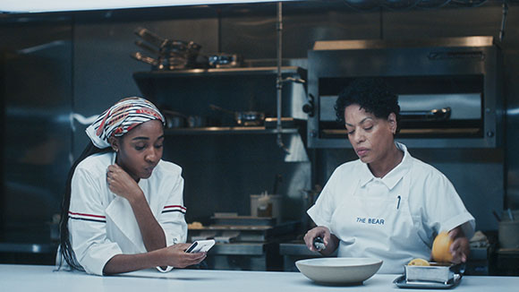 A young chef looks at a phone while an older chef prepares food in a stainless steel kitchen.