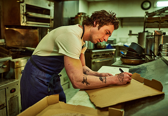 A man in a white t-shirt and blue apron writes on a cardboard box in a commercial kitchen.