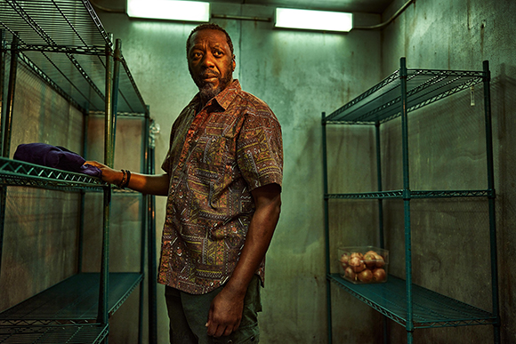 A man in a patterned shirt stands between two large green metal wire shelving units in a dimly lit room.