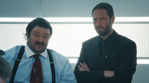 A large man in a blue striped shirt and tie stands next to a man in a dark suit with arms crossed.