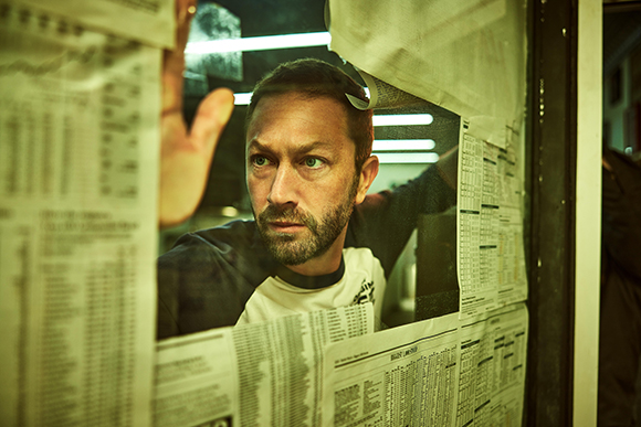 A man with a beard peers through a glass window covered in various printed documents and lists.