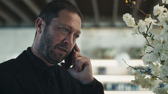 A man with short hair and a beard wearing a black suit talks on a mobile phone near white flowers.