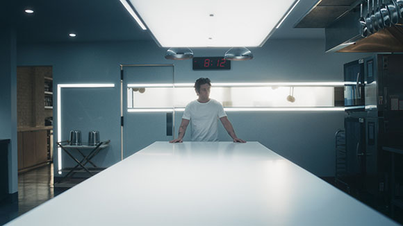 A man in a white t-shirt stands behind a long, illuminated white counter in a dark, modern kitchen.