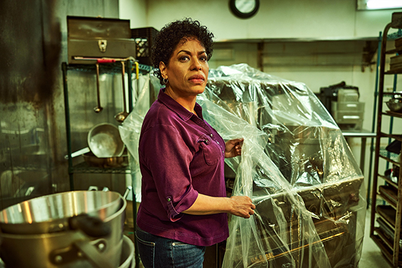 A woman in a purple shirt covers large kitchen equipment with clear plastic sheeting in a commercial kitchen.