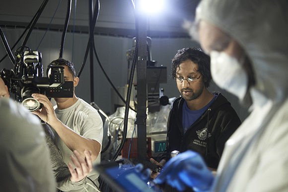 A camera operator films a subject while a crew member in a mask and hood looks at a device.