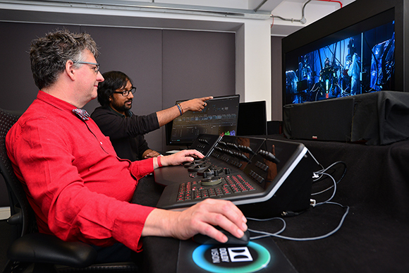 Two men sit at a control console with monitors in a dark studio environment.