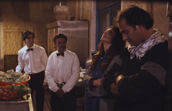 Two men in white shirts and bowties stand near a woman and a man in a dark sweater.