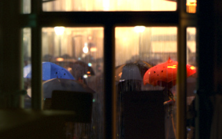 Blurred view through a window of people walking with colorful umbrellas in the rain at night.