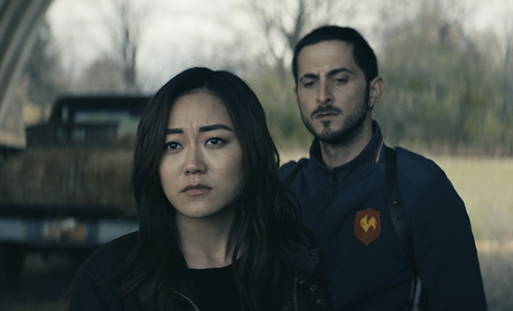 A woman with long dark hair and a man with a beard look forward with somber expressions outdoors.