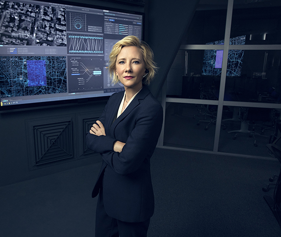A woman in a dark suit stands with arms crossed in a high-tech command center with digital displays.