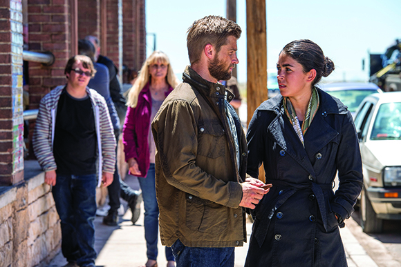 A man in a brown jacket and a woman in a black trench coat talk on a sunny sidewalk.