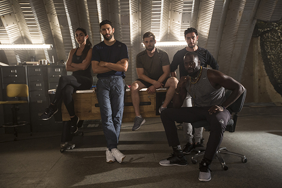 Five people in athletic and casual wear pose in a large, industrial room with ribbed metal walls.