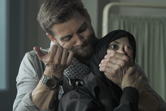 A man with a beard holds a woman wearing a black hijab, covering her mouth with his hand.