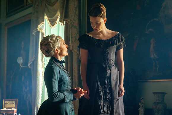A woman in a dark blue suit looks up at a taller woman in a black gown in a vintage room.