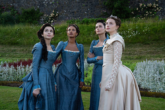 Four women in elegant 18th-century gowns stand in a lush green garden, looking upward.