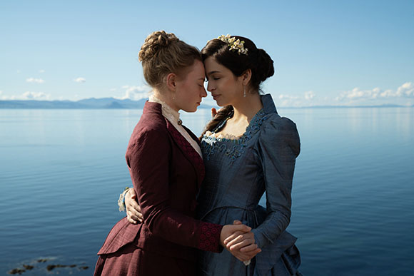 Two women in historical gowns stand close together, foreheads touching, against a calm blue ocean.