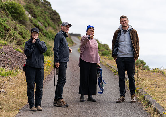 Four people stand on a narrow paved path winding through a grassy, hilly landscape under an overcast sky.