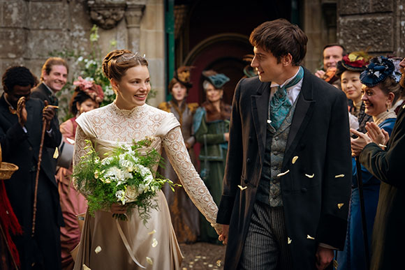 A bride in a lace gown and a groom in a black coat walk hand-in-hand through falling flower petals.