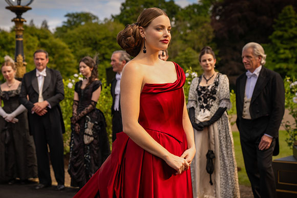 A woman in a red one-shoulder gown stands before a group of people in formal period clothing outdoors.