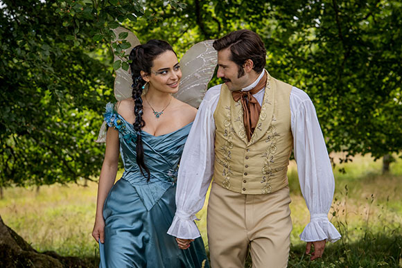 A woman with fairy wings in a blue dress and a man in a tan waistcoat hold hands in a grassy field.