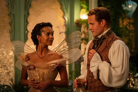 A woman wearing fairy wings and a gold dress looks at a man in a patterned vest and white shirt.