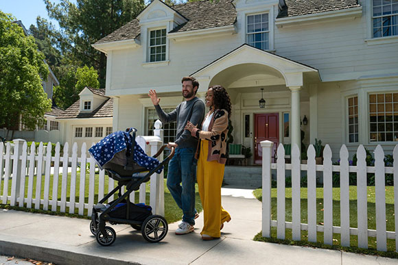 A man and woman walk a baby stroller down a sidewalk in front of a white house with a red door and a white picket fence.