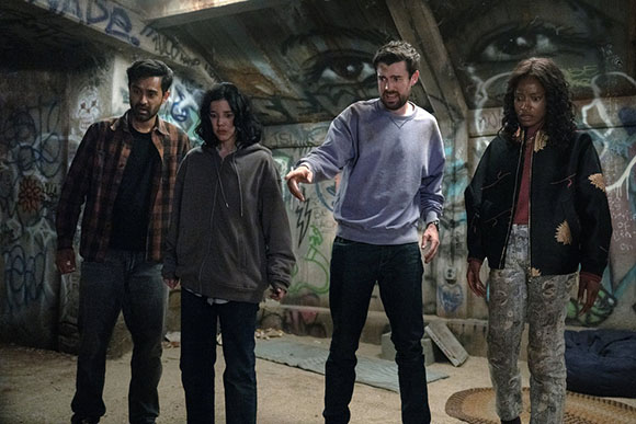 Four people, two men and two women, stand in a dimly lit room covered in graffiti. One man gestures with his hand.