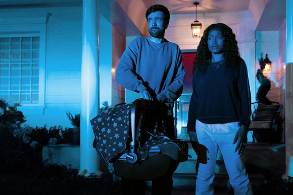 A man and a woman stand on a porch at night. The man holds a baby carrier with a star-patterned blanket. Blue light illuminat