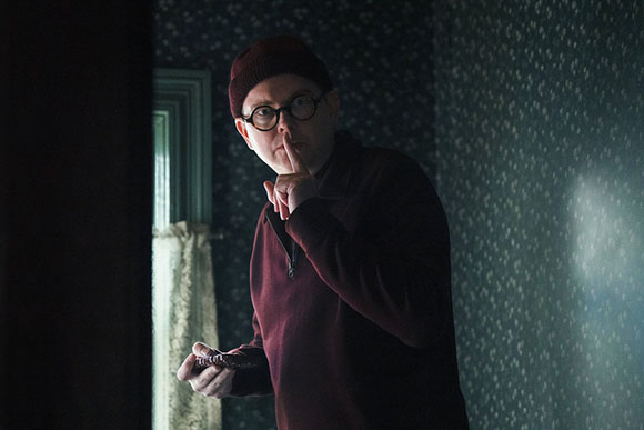 A man wearing a maroon beanie, round glasses, and a maroon sweater holds his finger to his lips in a shushing gesture.