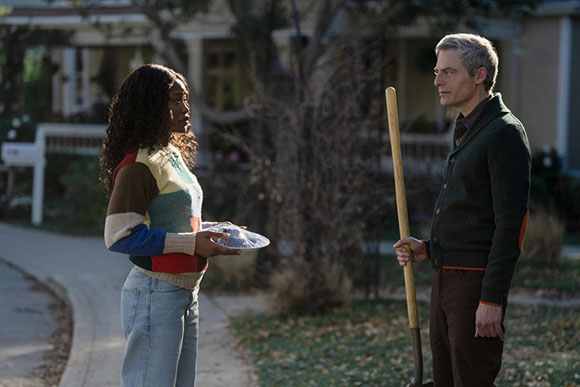 A woman with dark curly hair holds a blue object. A man with graying hair holds a shovel. They stand on a sidewalk in front o