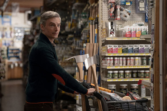 A man with graying hair wearing a dark green sweater with orange elbow patches looks over his shoulder while standing in an a