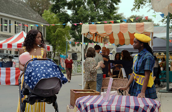 Two young women stand near stalls at an outdoor fair. One woman pushes a stroller and holds cotton candy, the other wears a y