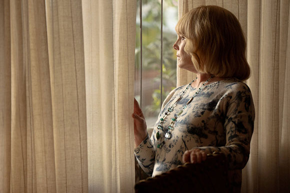 An older woman with blonde hair and a patterned top looks out a window, her hand resting on the curtain.