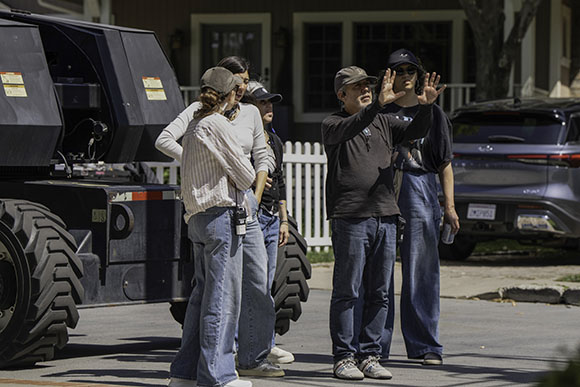 A film director gestures with his hands while directing a scene on a residential street. Several crew members stand nearby, a