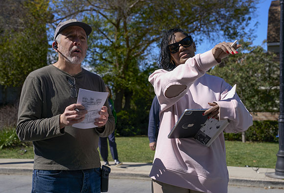 A man in a baseball cap holds papers and a pen, looking to the side. A woman in sunglasses and a pink sweatshirt points with